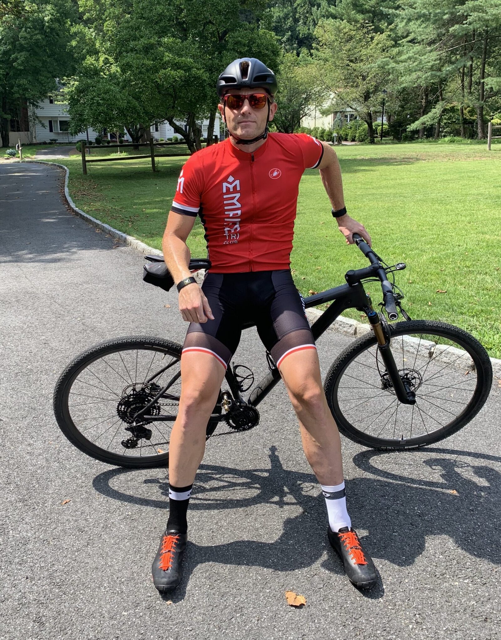 A man in red shirt and black shorts standing next to bicycle.